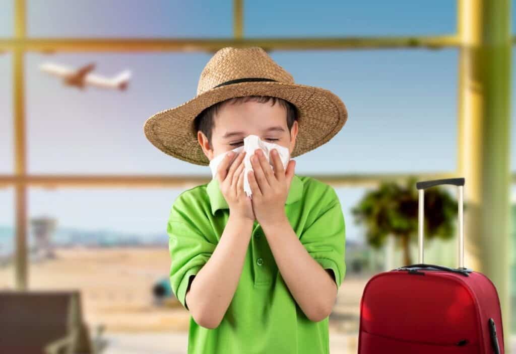 A young boy in a green shirt and straw hat, sick on holiday, blows his nose with a tissue at an airport, with a red suitcase and a plane visible in the background.